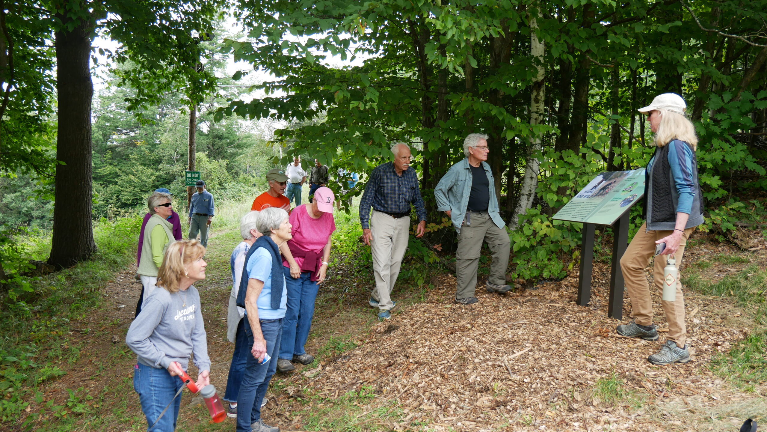 A woman speaking to a group of people in front of a trail sign