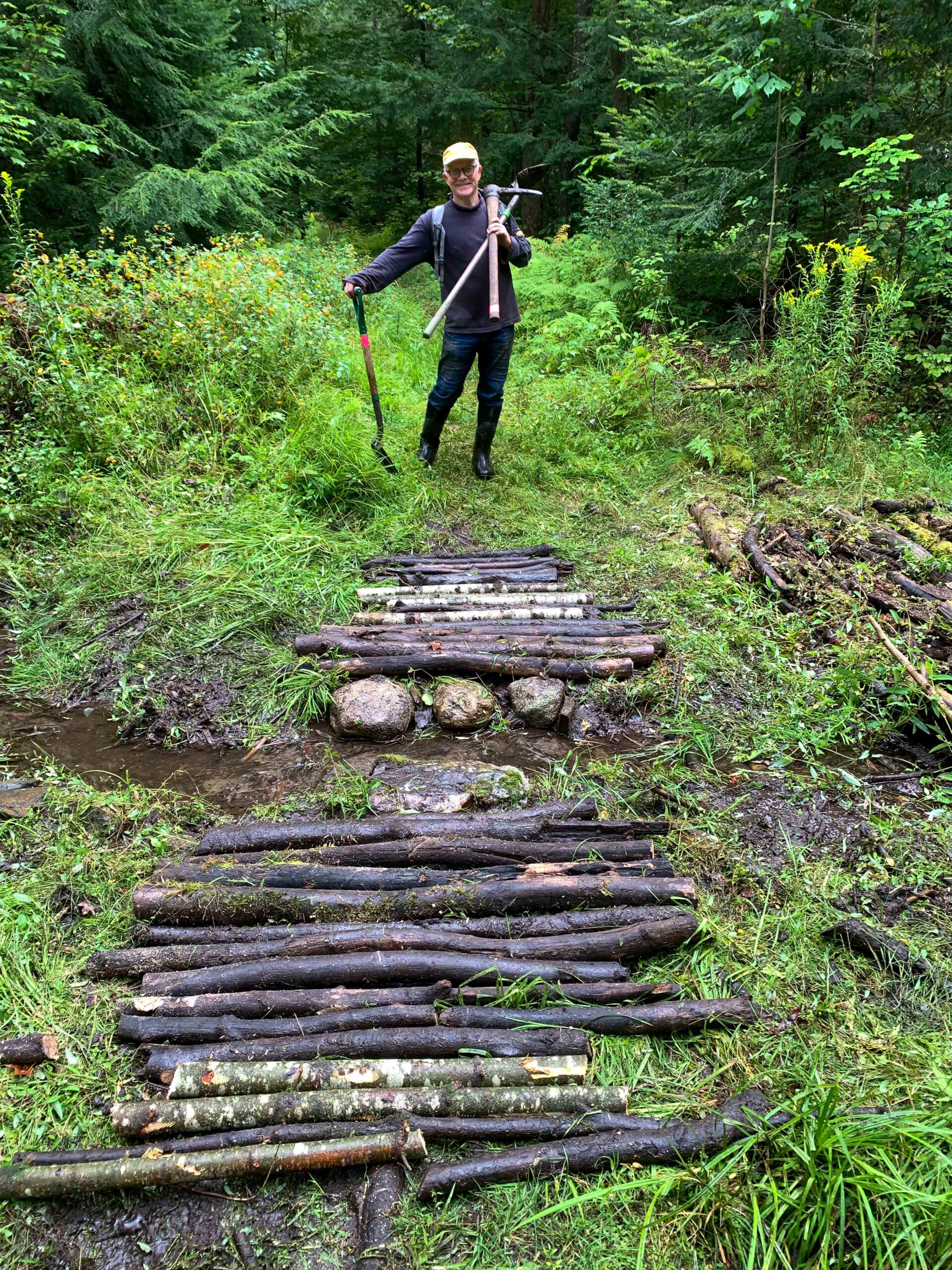 A person holding trail tools standing in front of a bridge