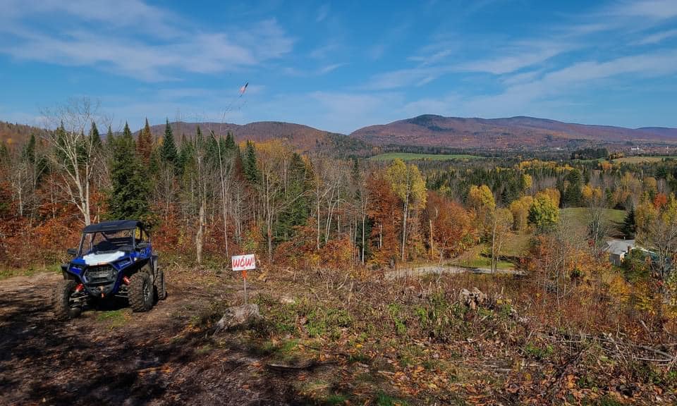 Blue ATV parked on a dirt path beside a "WOW" sign in a scenic landscape with autumn trees and distant mountains under a clear sky.