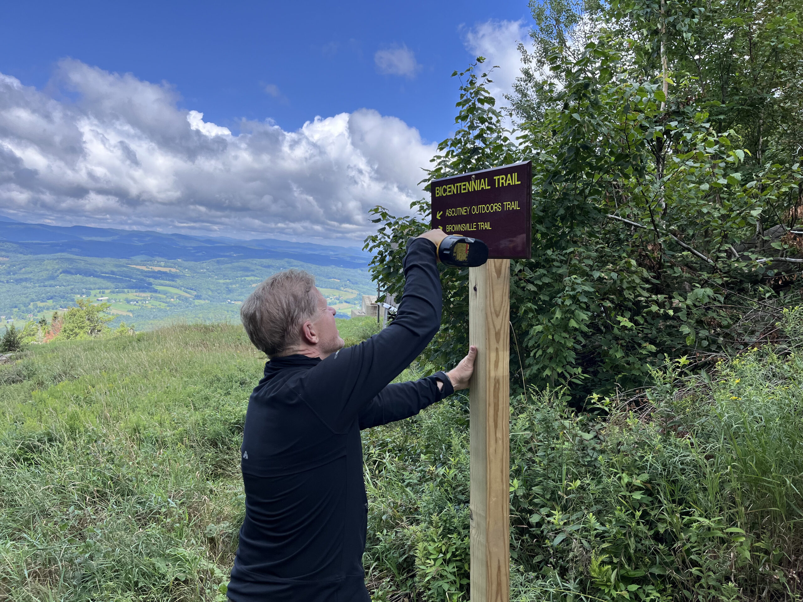 A person is attaching a Bicentennial Trail sign to a wooden post on a hillside with a view of hills and a cloudy sky in the background.