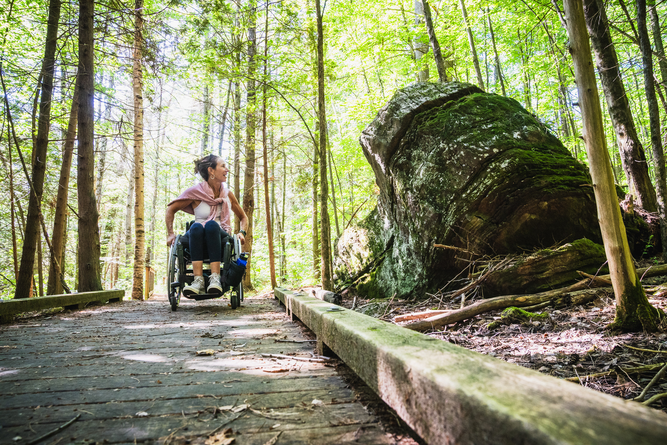 woman in wheelchair on boardwalk in woods admiring large glacial erratic
