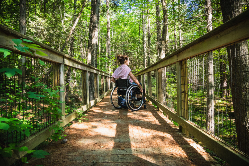 woman in wheelchair on boardwalk with side rails going through woods